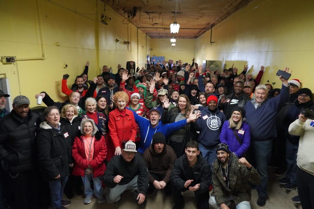 Large group posing indoors in a hall, many wearing red and blue jackets, some raising their arms.