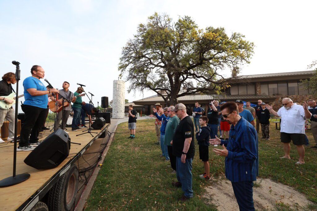 People standing in a grassy outdoor area near a stage, listening to musicians performing.