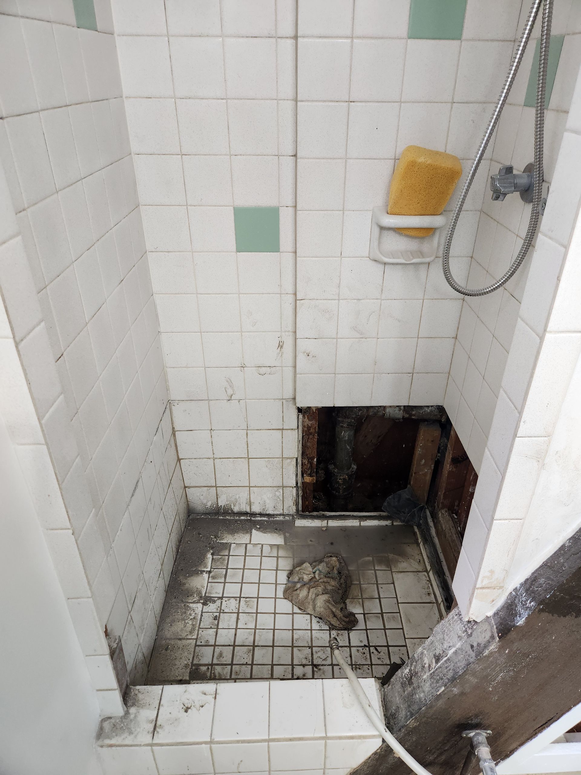 A damaged shower stall with missing tiles and a hole in the floor. A sponge sits on a soap dish.