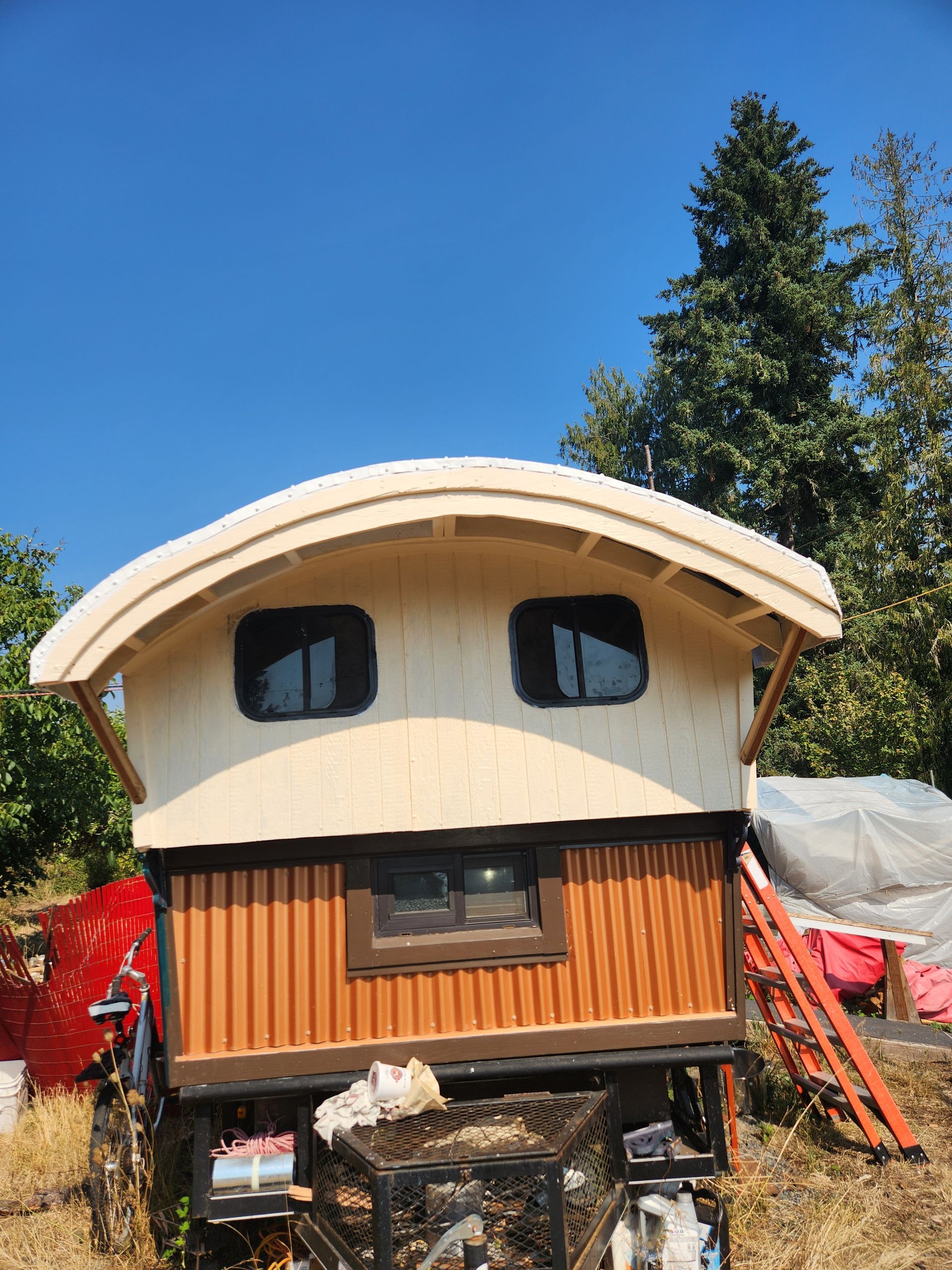 Wooden wagon-like structure with a curved roof, windows, and brown corrugated siding, set outdoors under a blue sky.