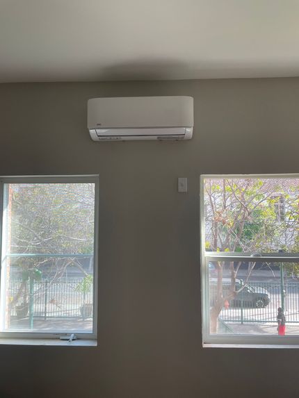 A white wall-mounted air conditioning unit above two windows. The windows face outdoors with trees visible.