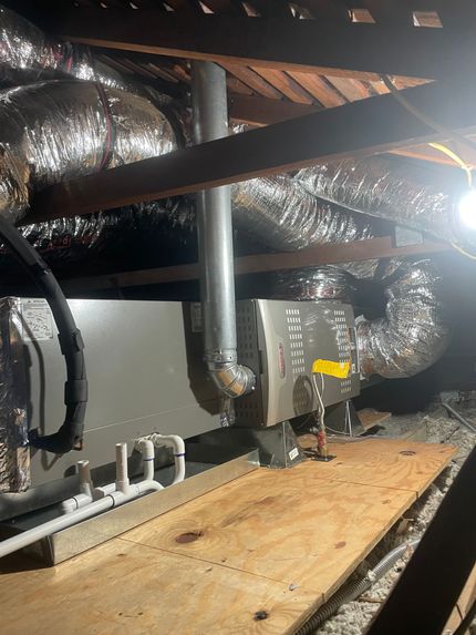 HVAC unit in an attic.  Silver ductwork and a metal flue extend from the unit.  Wooden beams and insulation are visible.