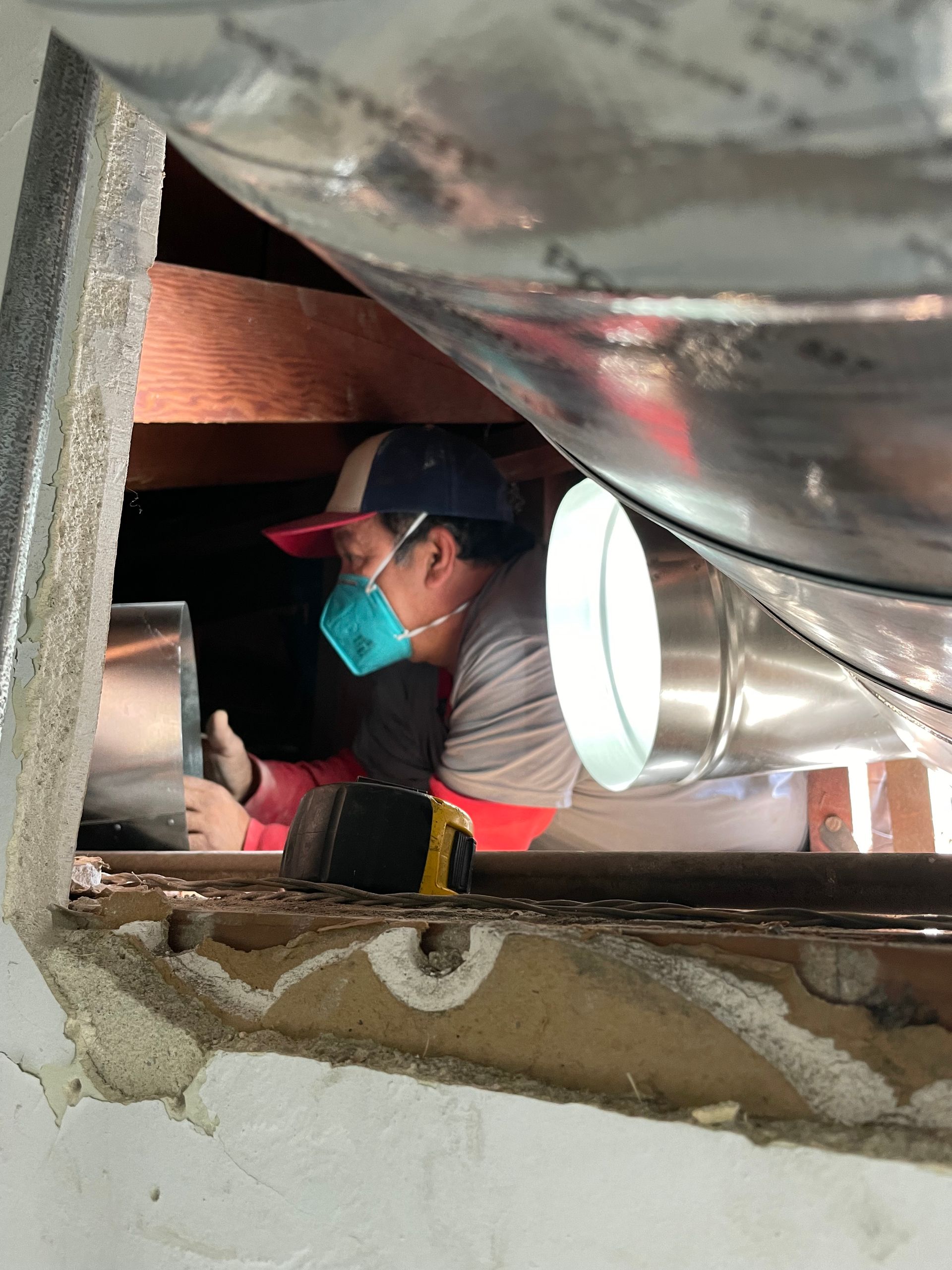 A person in a cap and mask works on ductwork in an enclosed space. They are attaching a metal duct to a larger silver duct.