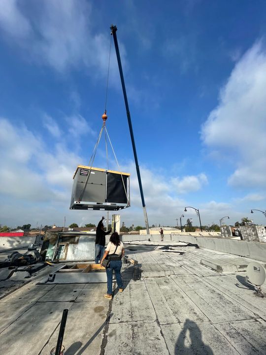 A crane lifts an HVAC unit onto a building's flat roof. Two people stand nearby, observing. Blue sky with scattered clouds.