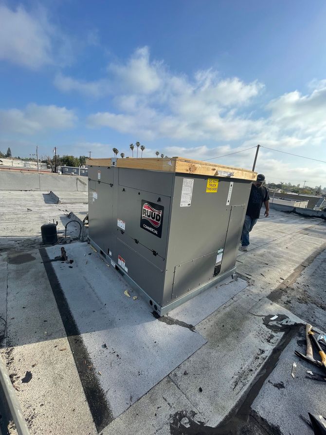 HVAC unit on a rooftop with a worker. The unit is gray with black accents, sitting on a gray roof with a blue sky background.