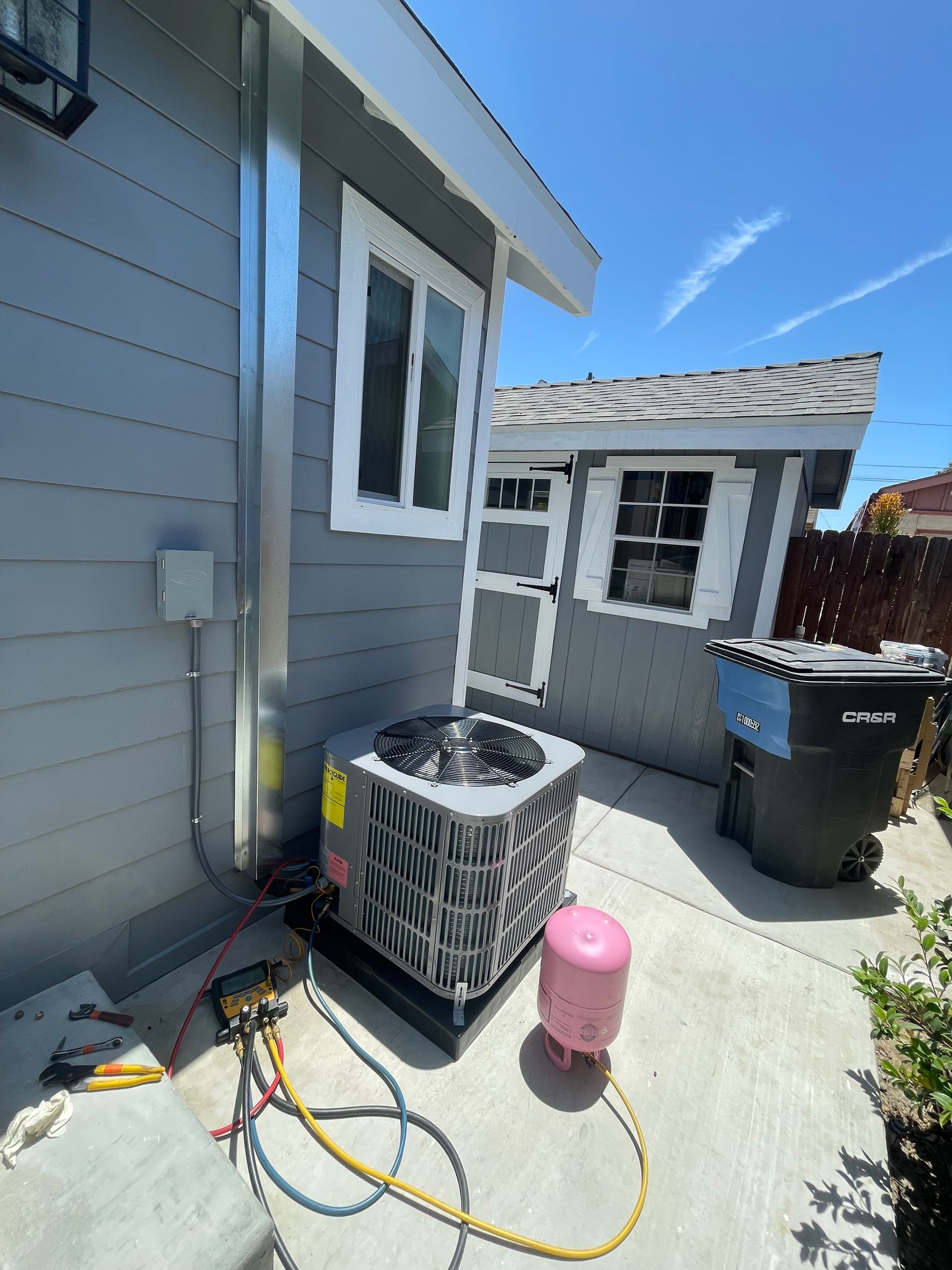 An air conditioning unit sits outside a gray house. It's connected to gauges and a pink tank; a trash bin and shed are nearby.