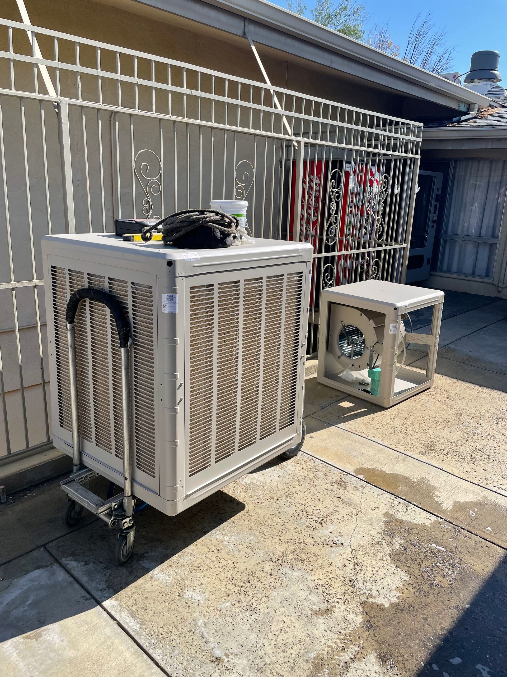Two evaporative coolers sit on a cracked concrete patio, against a fence and wall. The larger cooler has a handle and cart.
