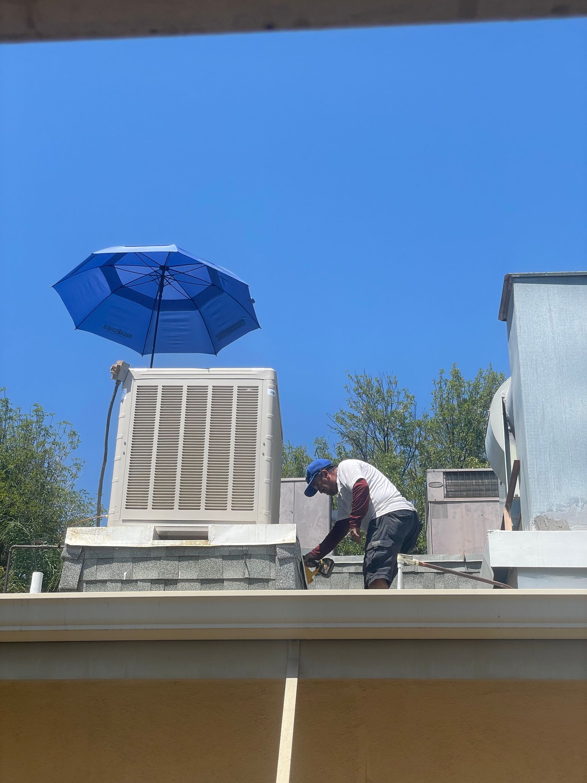 A person works on a rooftop unit under a blue umbrella on a sunny day. They are wearing a hat and gloves.