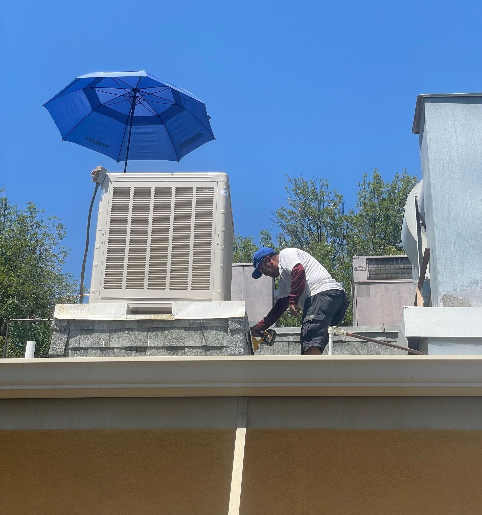 A worker on a rooftop near an air conditioning unit, using a tool under a blue umbrella on a sunny day.