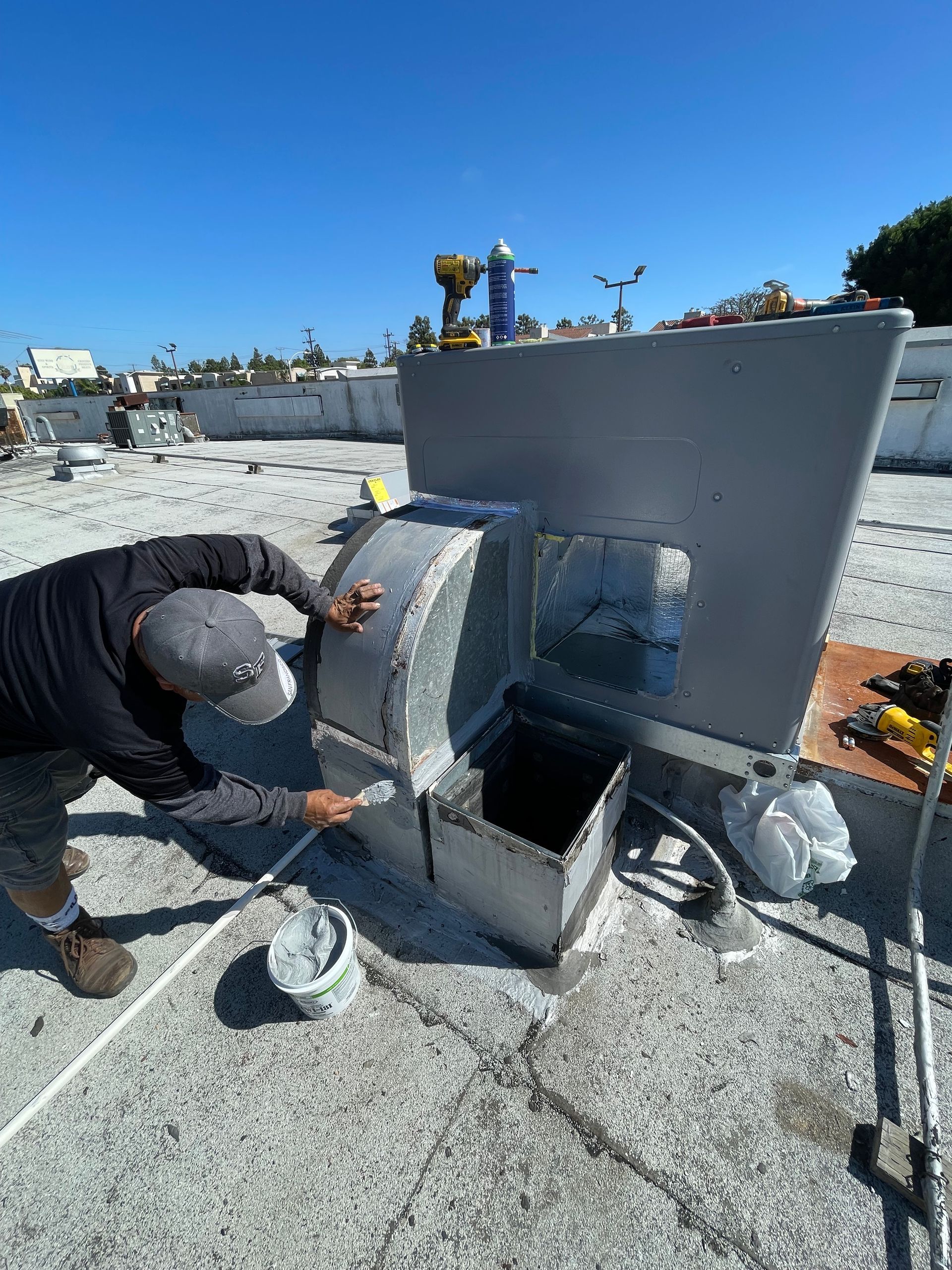 A worker repairs an HVAC unit on a rooftop, applying sealant with a brush. Bright blue sky overhead.
