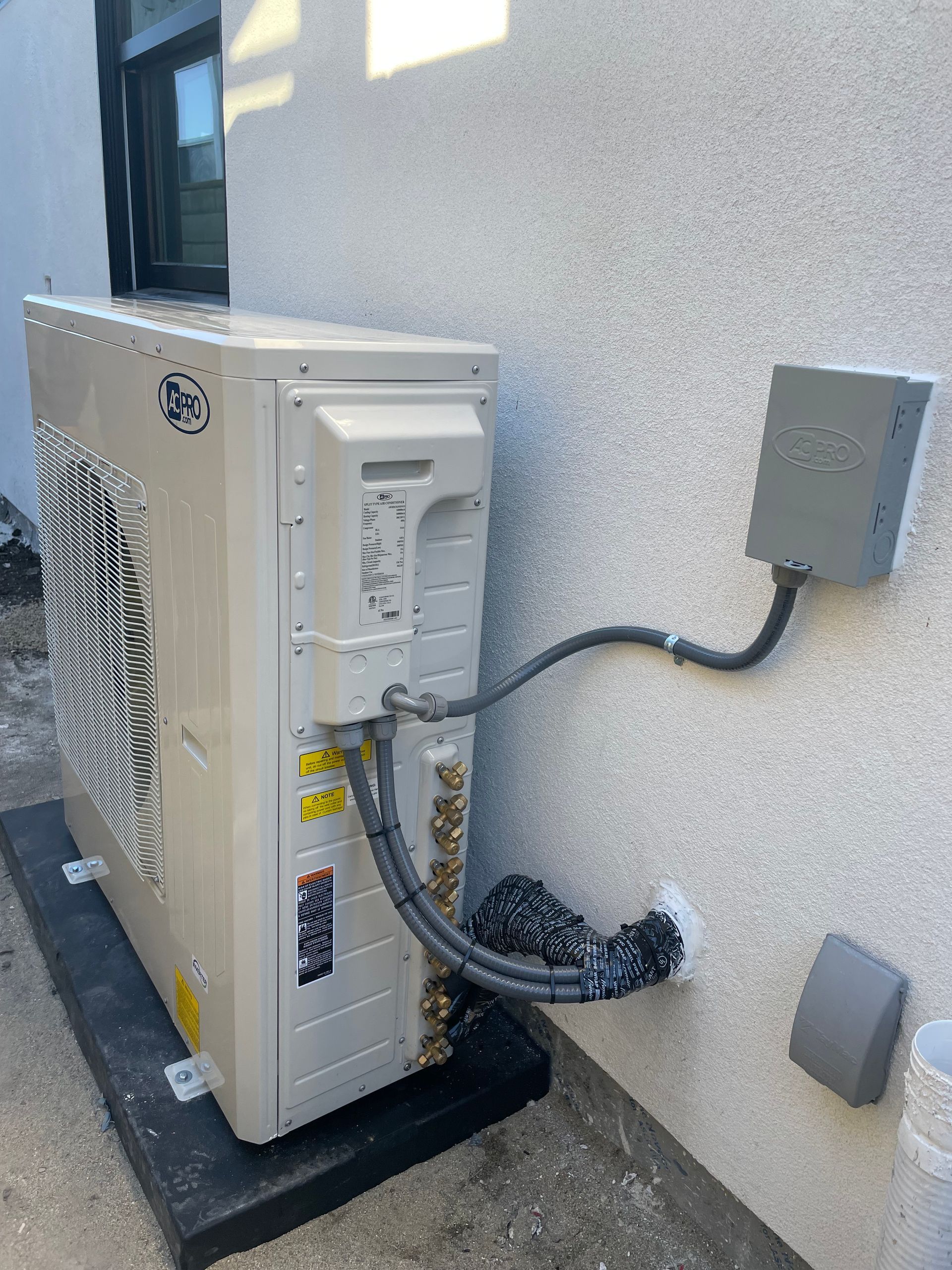 An outdoor HVAC unit mounted on a black pad against a light-colored stucco wall. Gray conduit connects to a nearby electrical box.