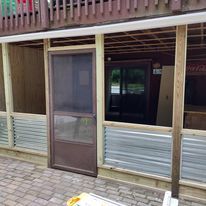 A porch with a screen door and a refrigerator underneath it.