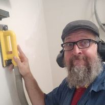 A man with a beard and glasses is using a sander on a wall.