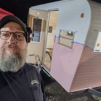 A man with a beard and glasses is taking a selfie in front of a pink trailer.