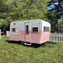 A pink and white trailer is parked in a grassy field next to a white picket fence.