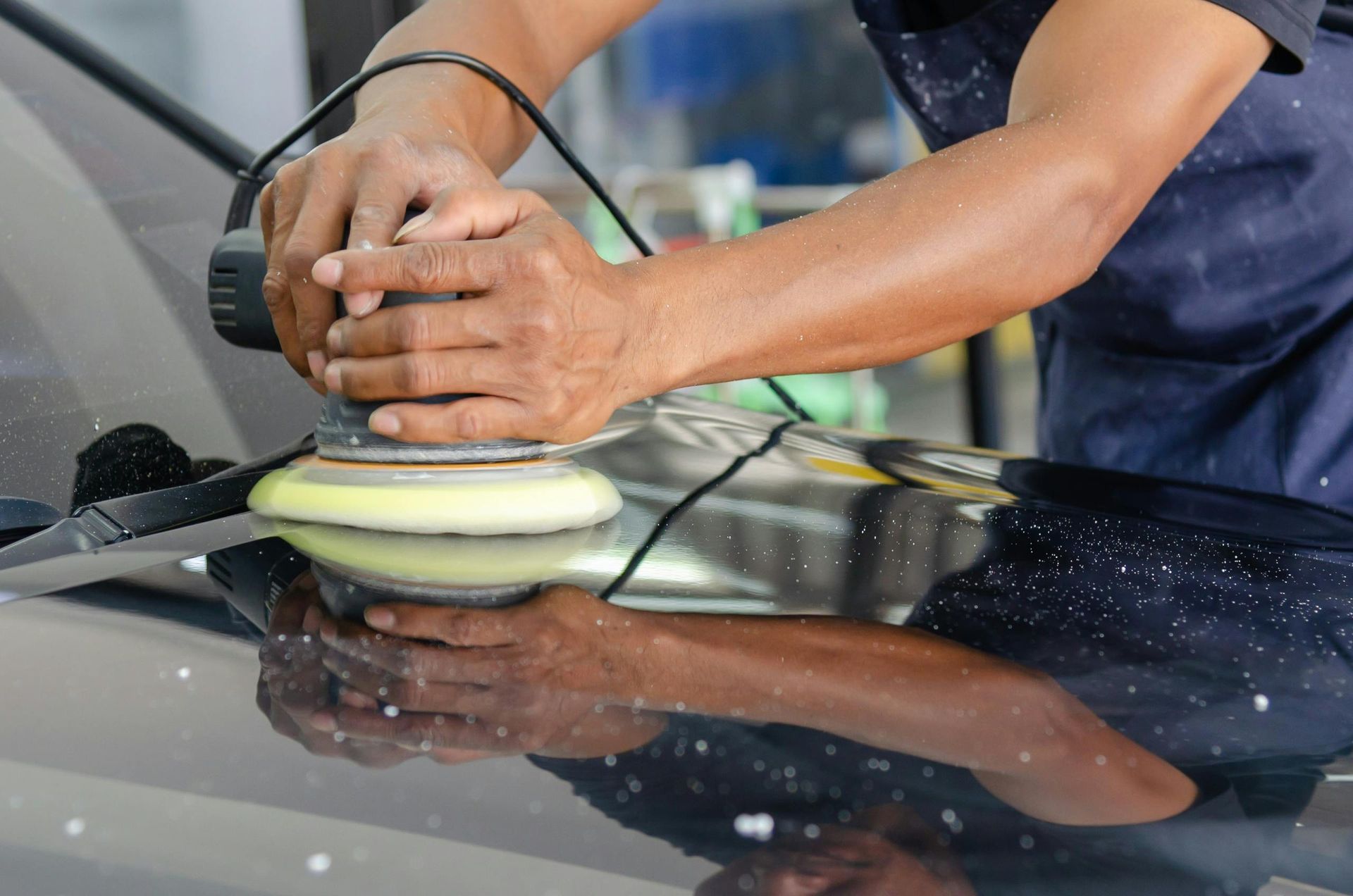 Person polishing a black car hood with a rotary buffer, reflecting in the glossy surface