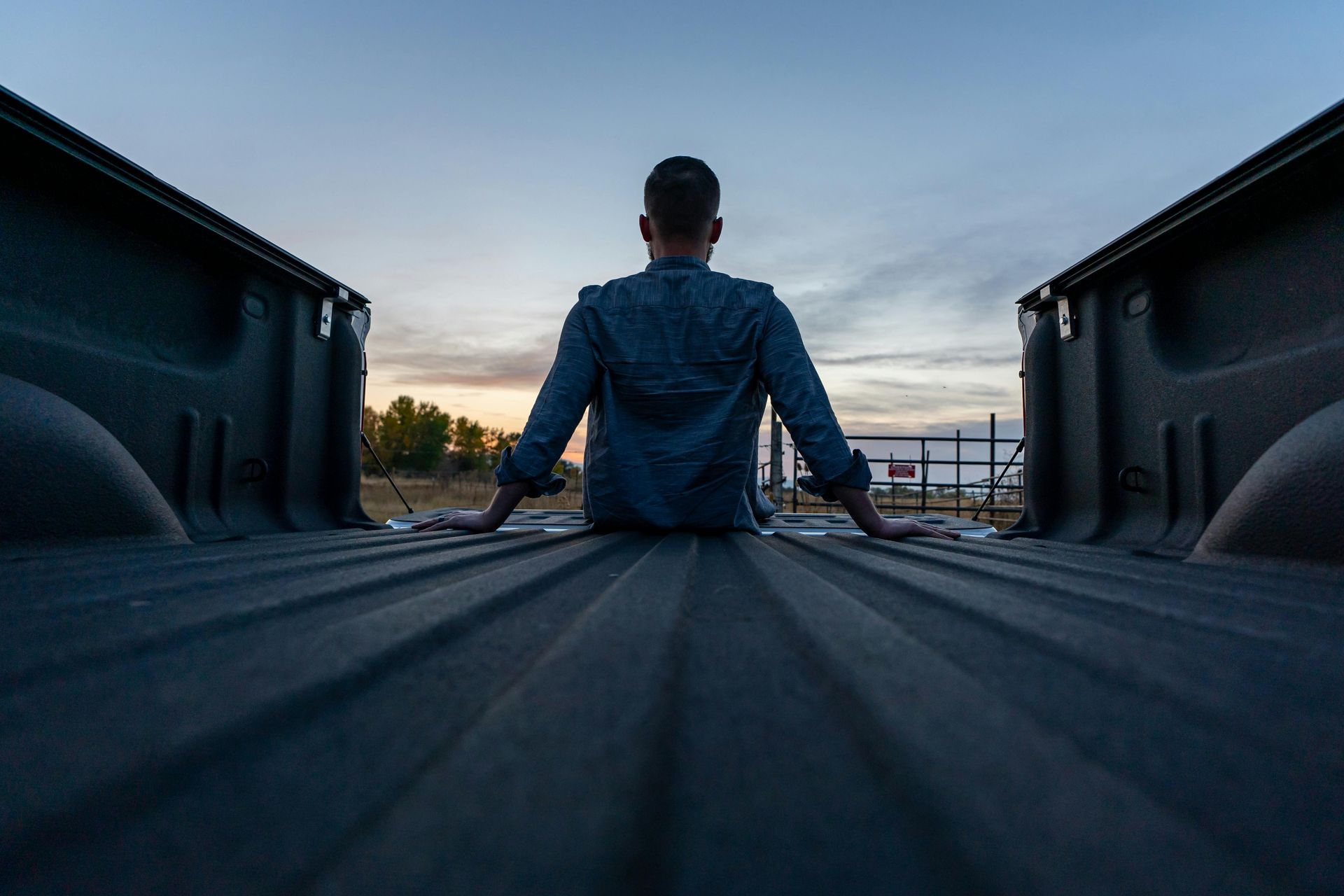 Person in a blue jacket sitting on a pickup truck tailgate at dusk, looking over a road.