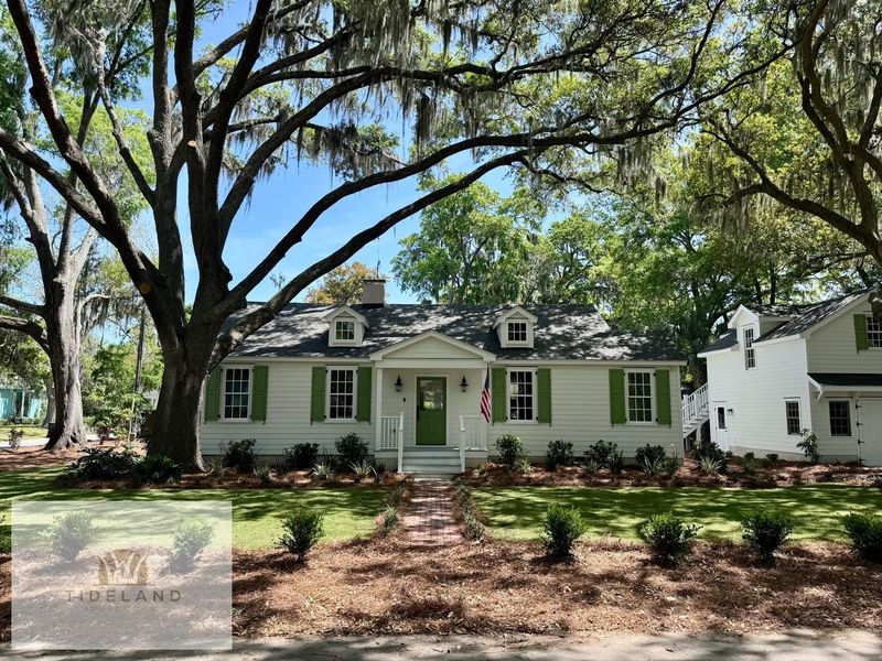 A white cottage-style house with green shutters, dormer windows, and a front porch, nestled under large oak trees.