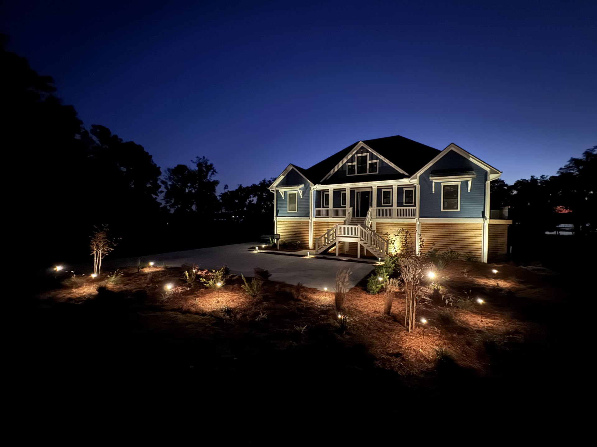 A blue house at night, illuminated by warm landscape lighting, with a light-colored porch, stairs, and a dark sky above.