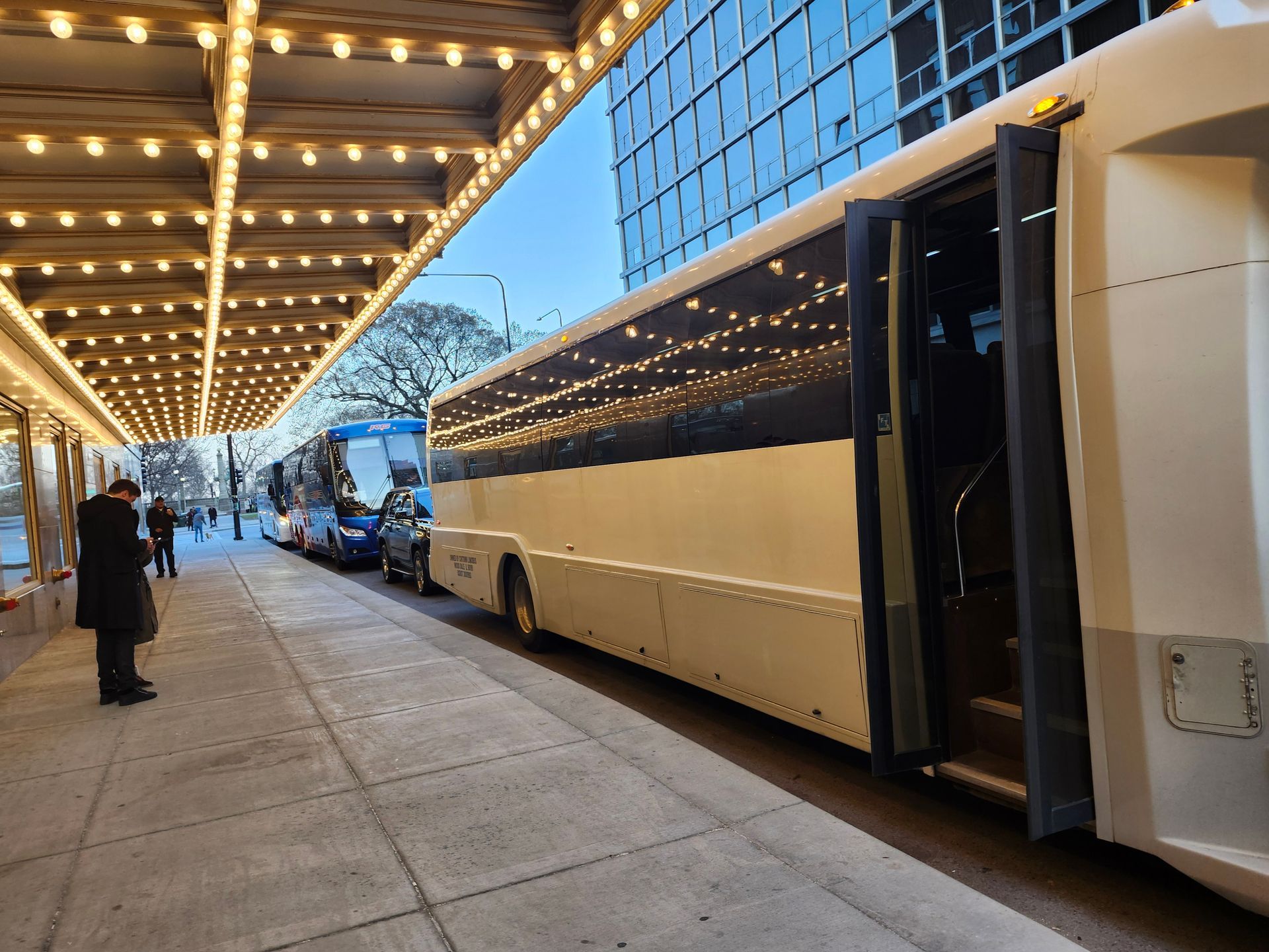 A large white bus with open door parked at a theater entrance under a canopy of lights.