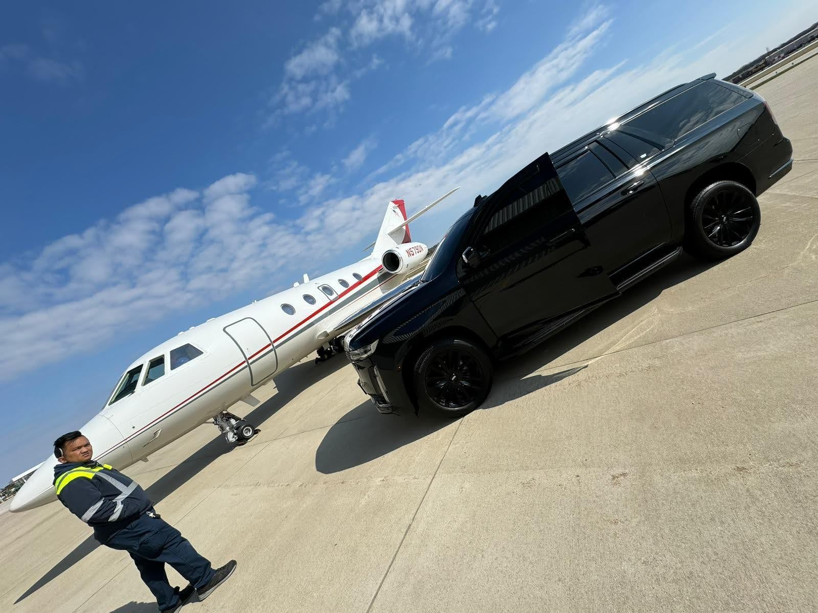 A black SUV and a private jet on a tarmac with a person in a work uniform, against a blue sky.