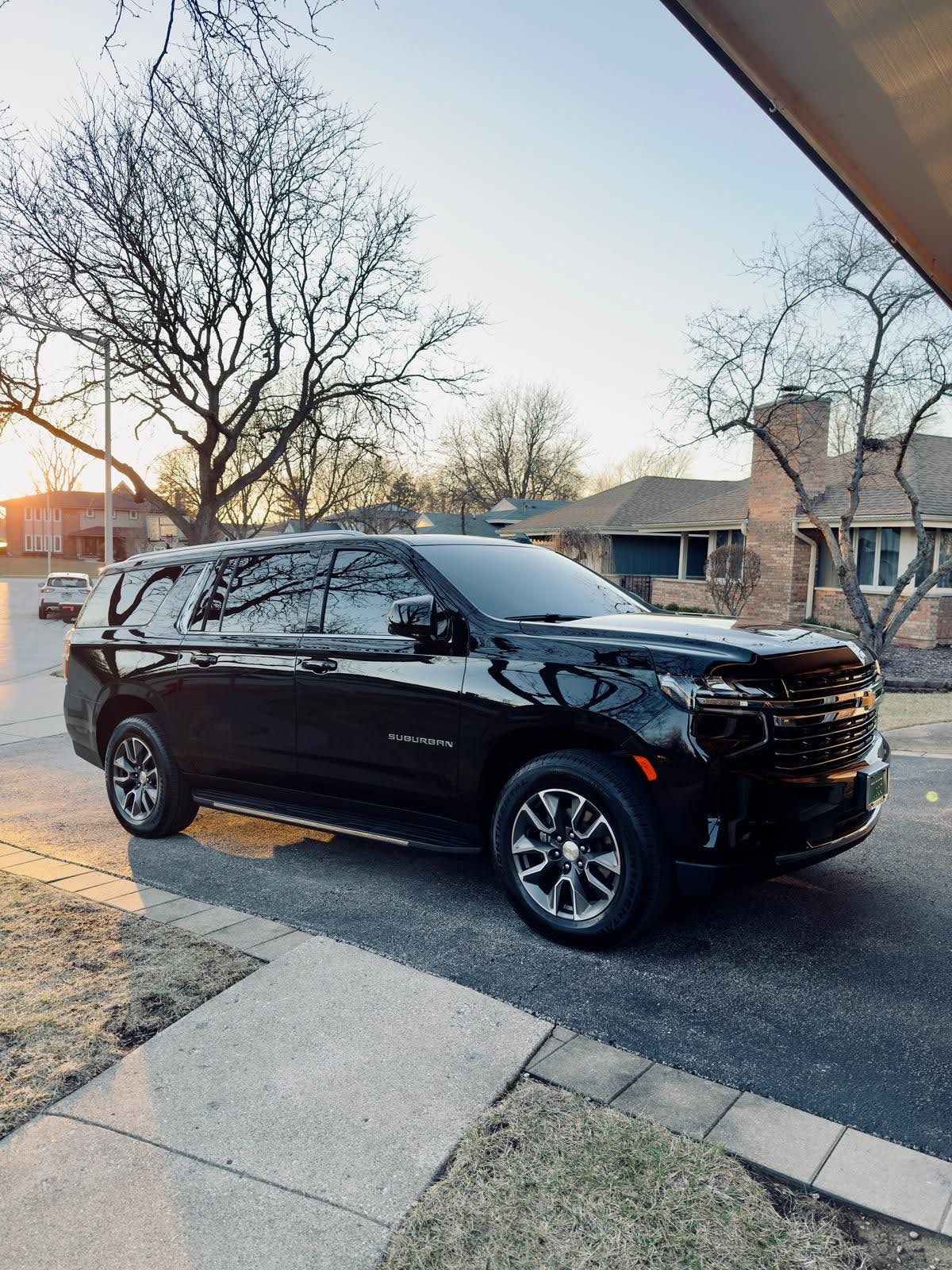 Black SUV parked on a sidewalk next to a residential house, with a tree in the background.