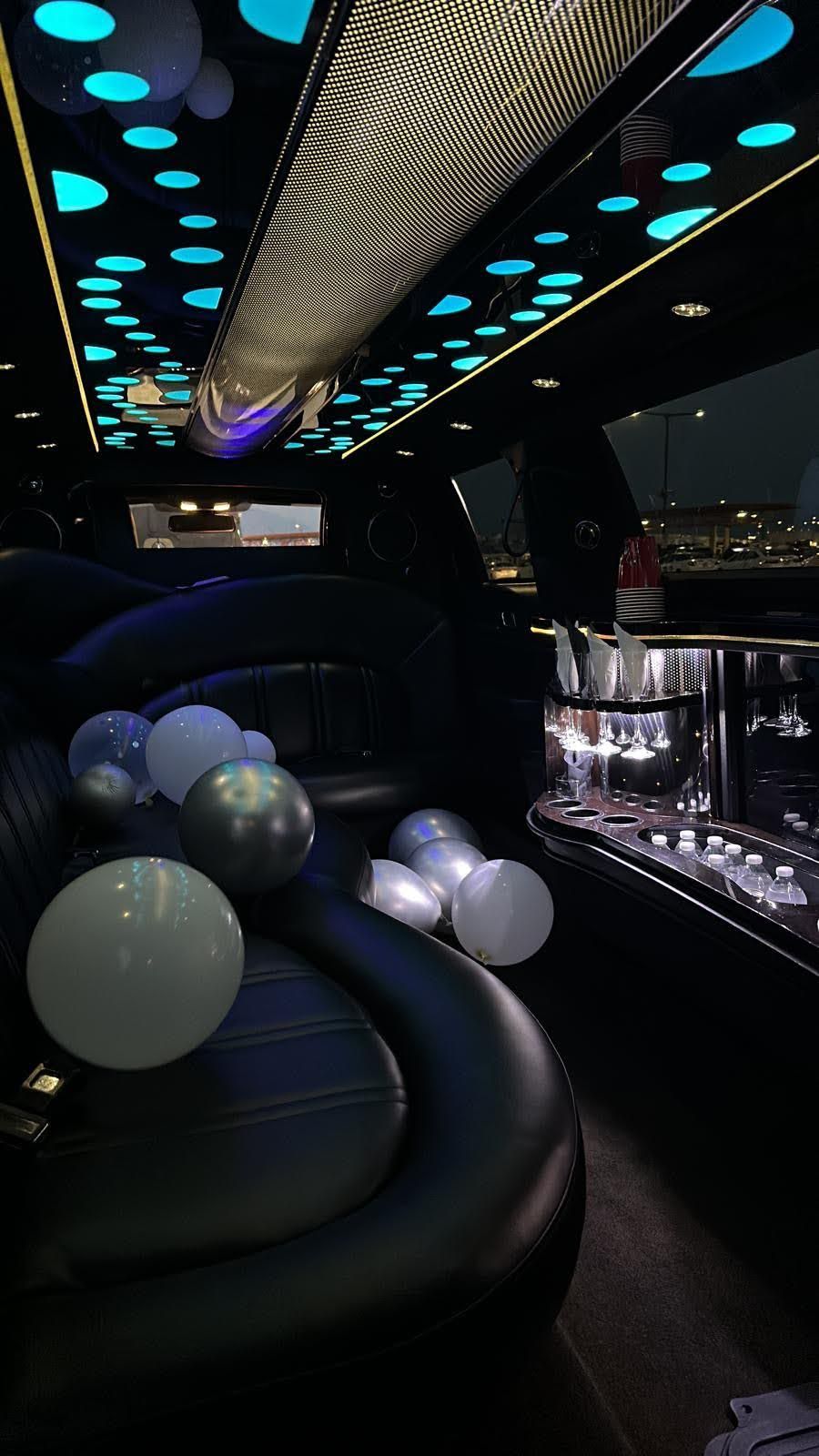Interior of a limousine decorated with balloons. The ceiling has blue lights. There's a bar area.