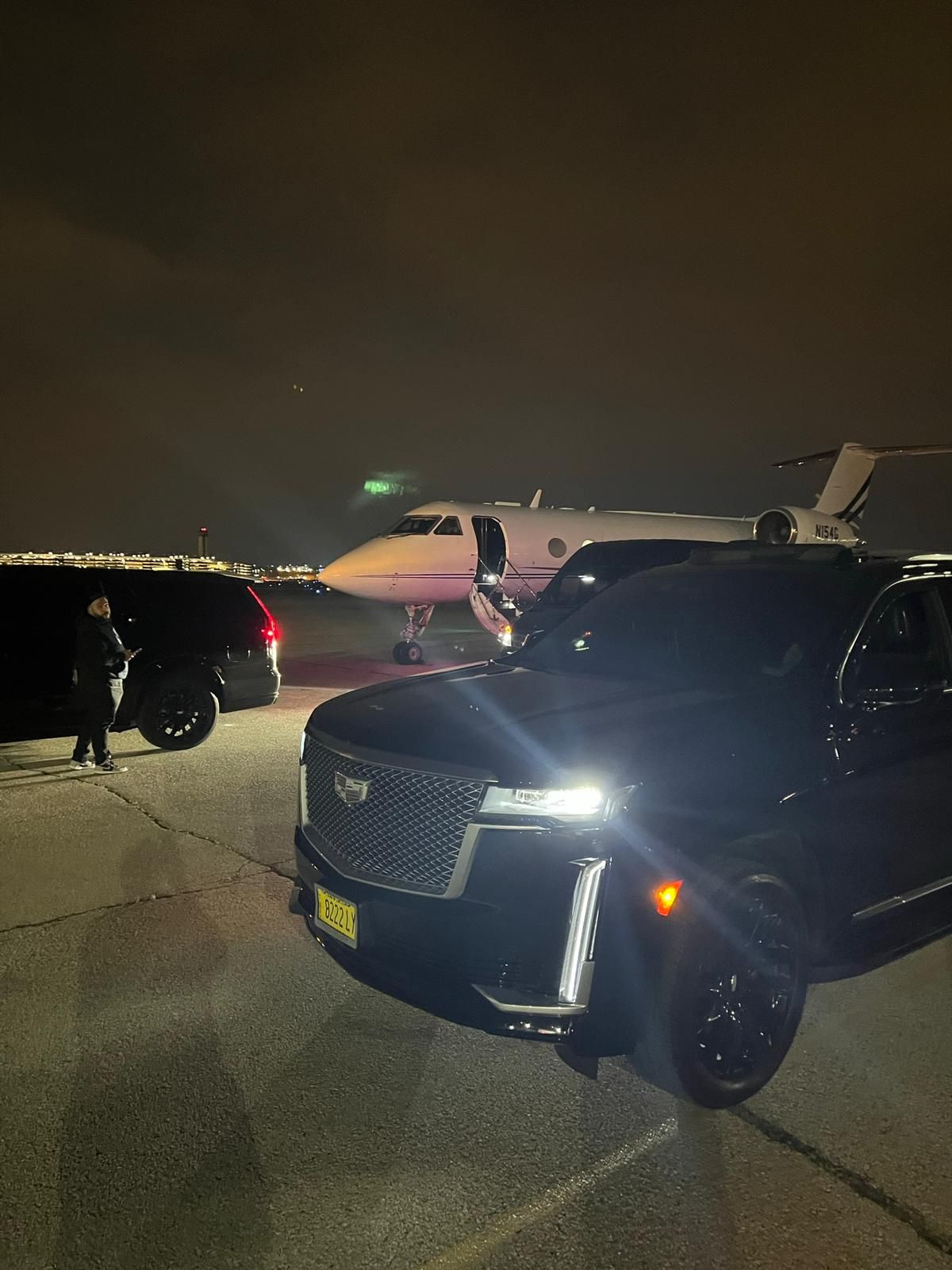 Cadillac Escalade with private jet at night; driver near jet, dark vehicles.
