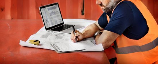 Worker in orange safety vest writing on papers beside a laptop on a red surface