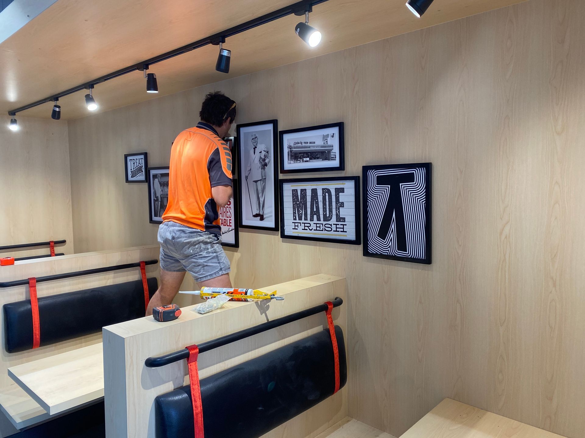 Man hanging framed artwork on a wood-paneled wall above booth seating, lit by track lighting.