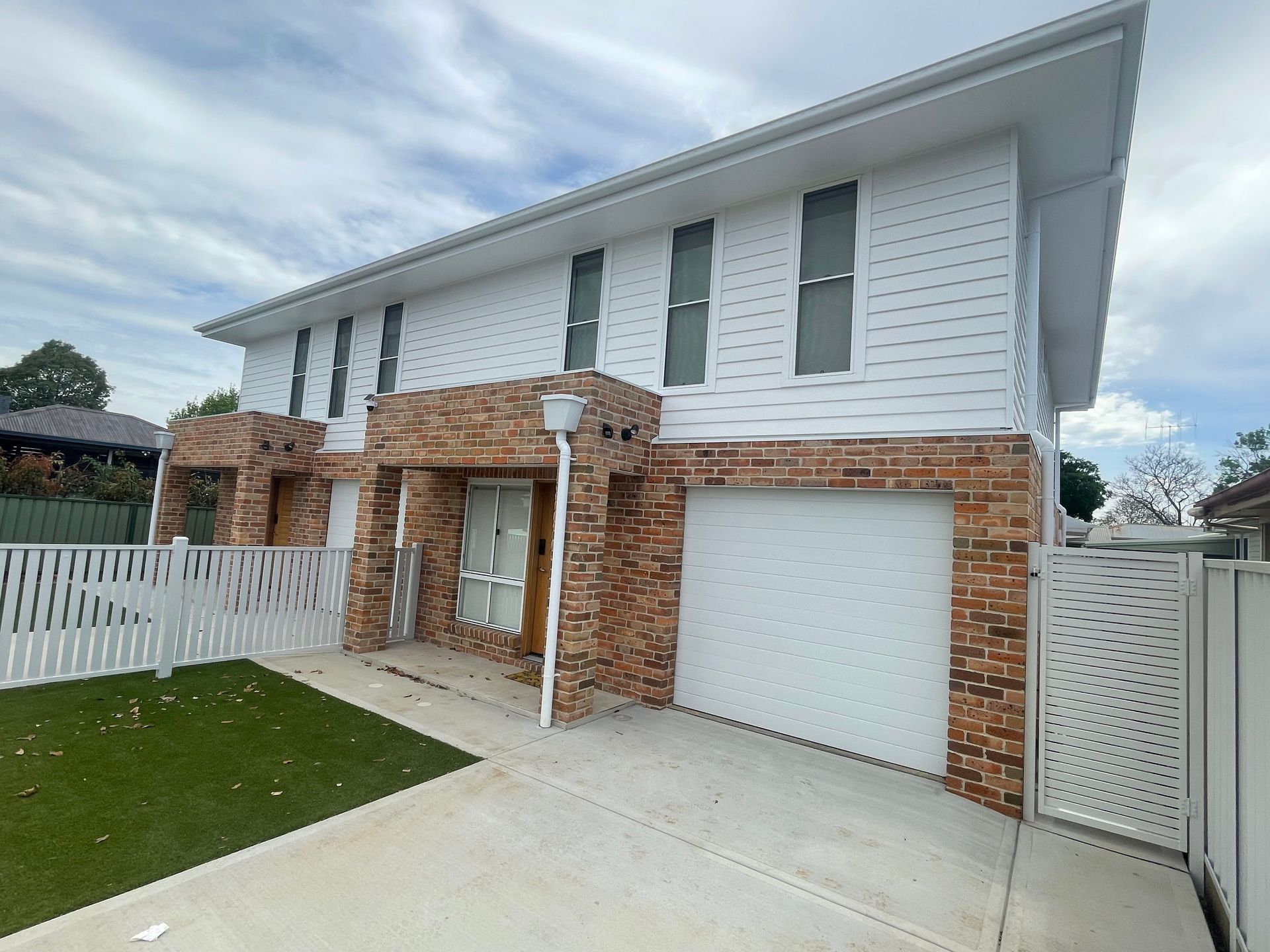 Two-story duplex with white siding, brick accents, garage doors, and white fencing under a cloudy sky.