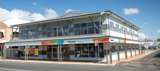 Two-story white building with glass windows, rainbow-colored sign, and a street in front.