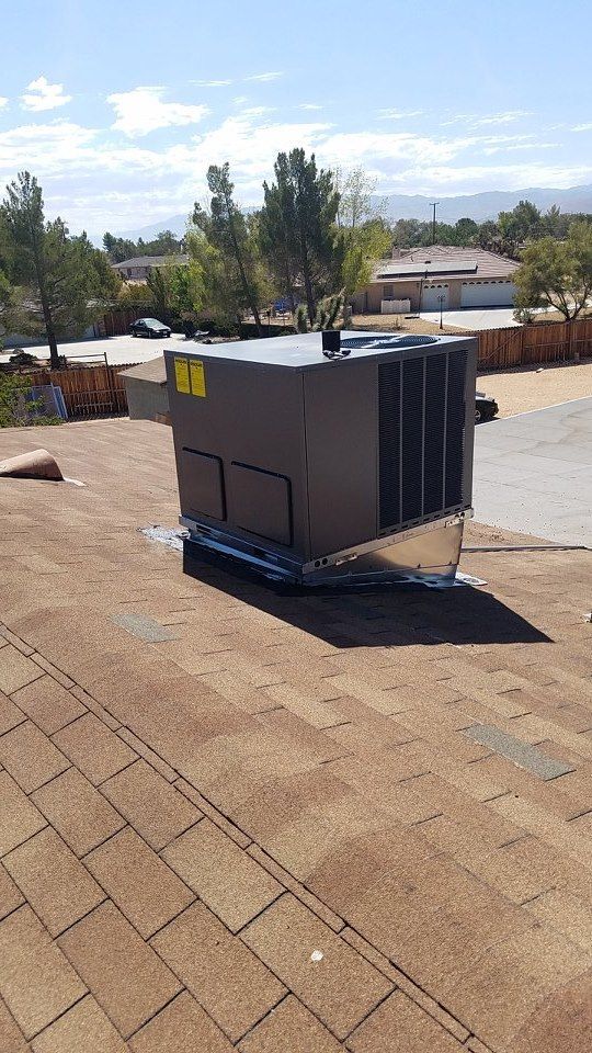 A large, gray HVAC unit sits on a brown shingle roof under a blue sky, near trees and houses.