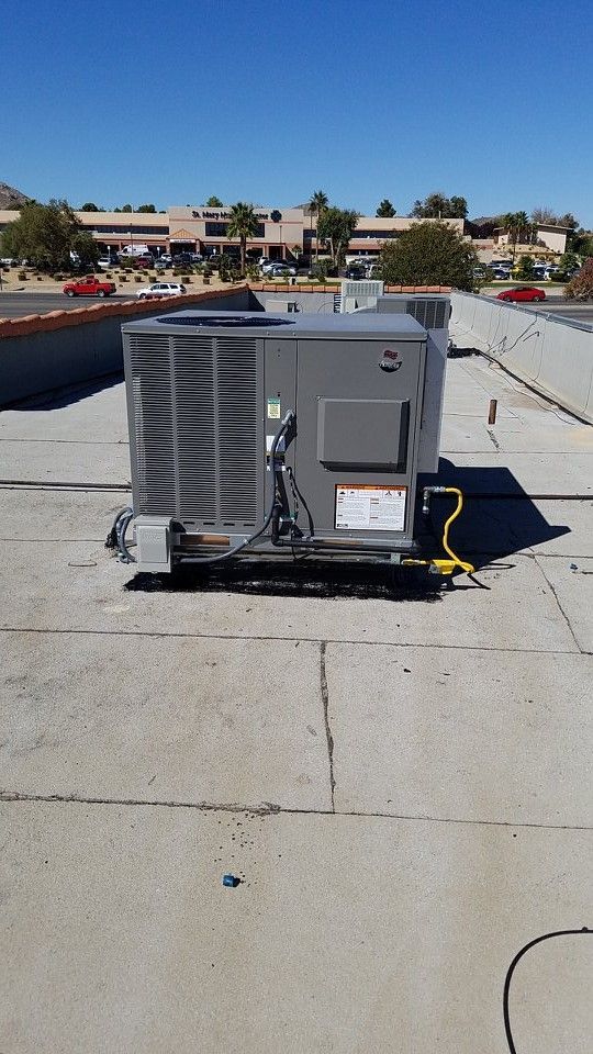 HVAC unit on a flat roof. Gray unit with vents, connected by yellow tubing. Sunny day, buildings in background.