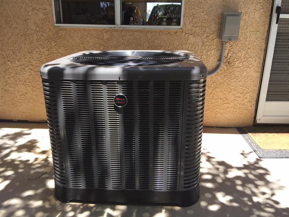 Dark-colored air conditioning unit outside a stucco building, with window and door visible.