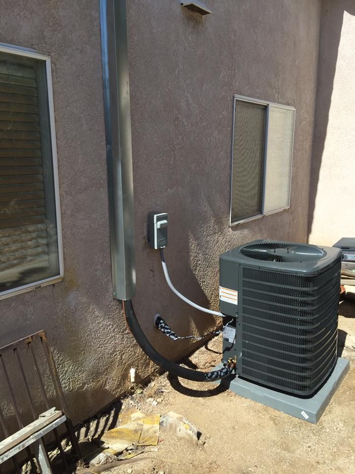 An air conditioning unit next to a brown stucco wall with electrical conduit and windows.