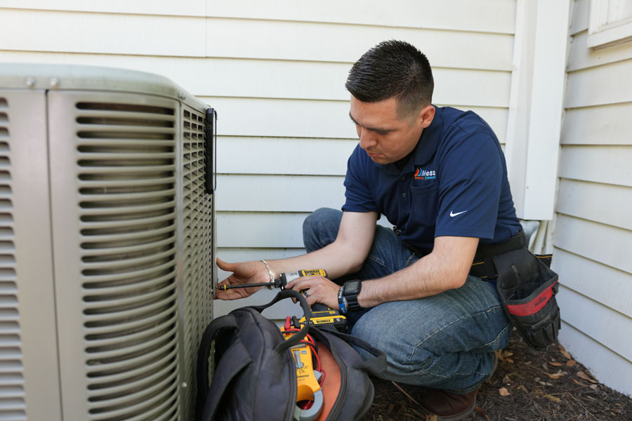 HVAC technician using a drill on an air conditioning unit outside a white building.