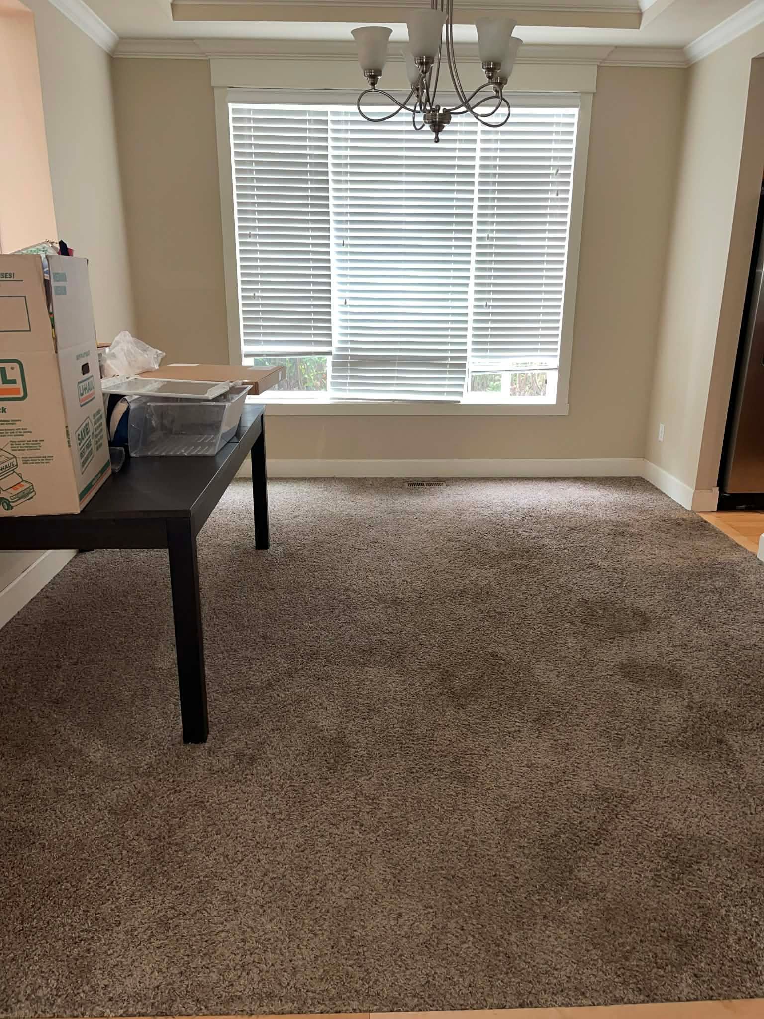 Empty carpeted room with a large window, chandelier, and a black table on the left.