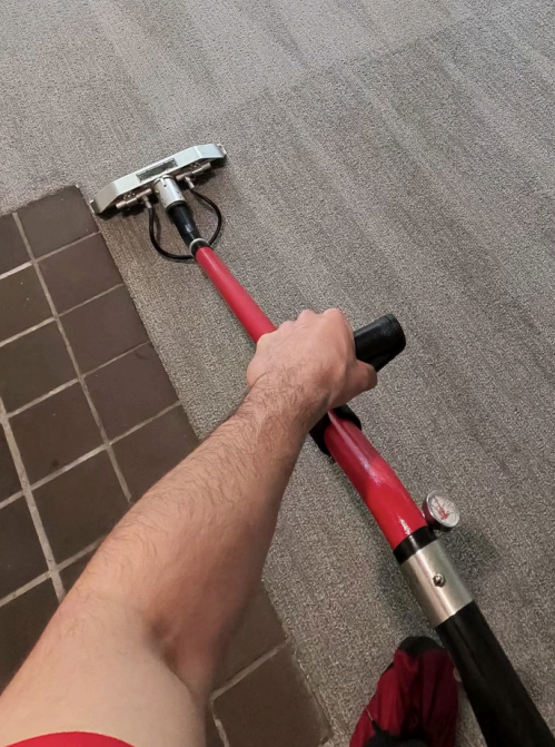 Hand holding a red pole squeegee cleaning a carpet beside brown floor tiles