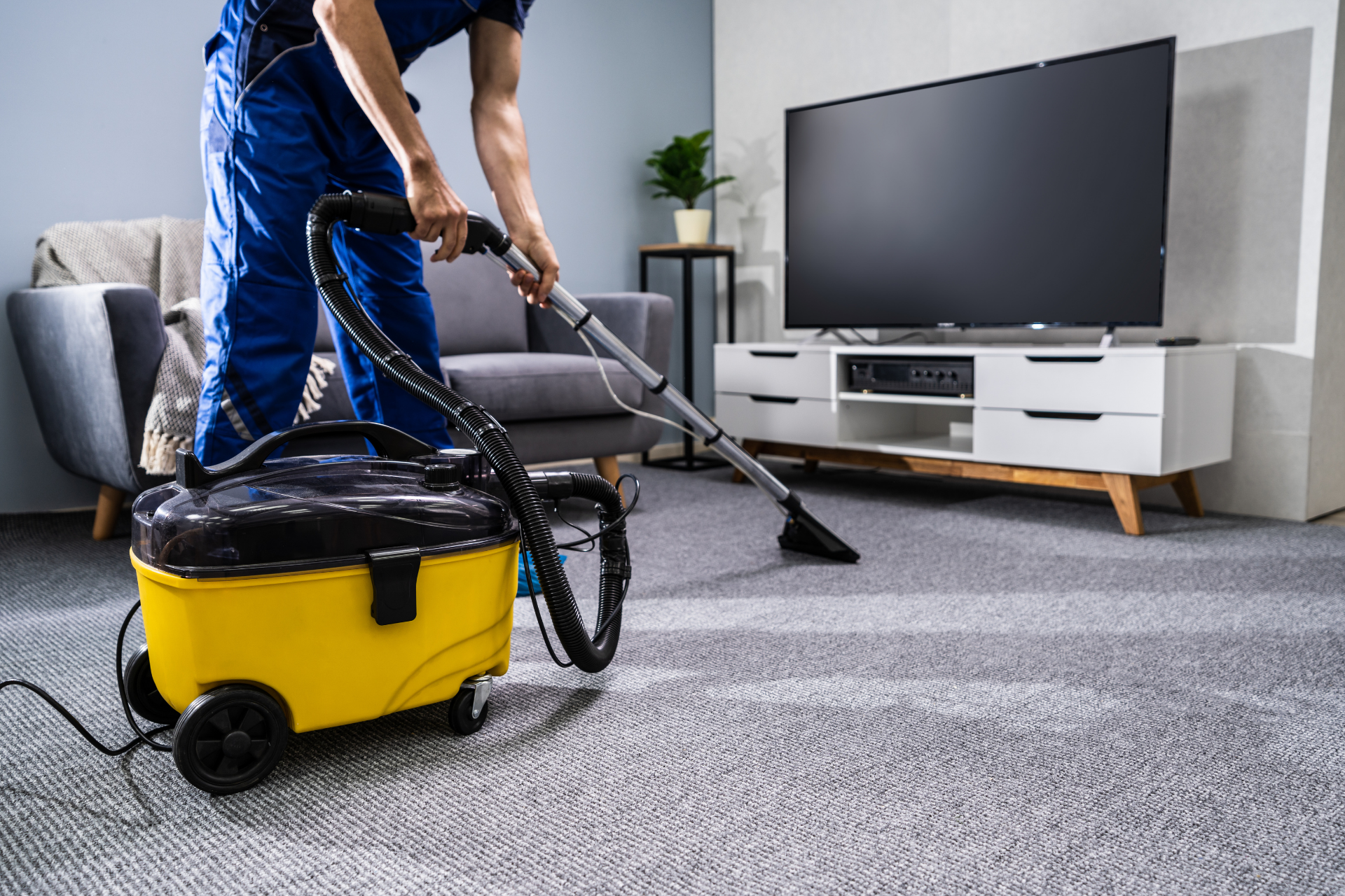 Person vacuuming a gray carpet in a modern living room with a yellow canister vacuum.