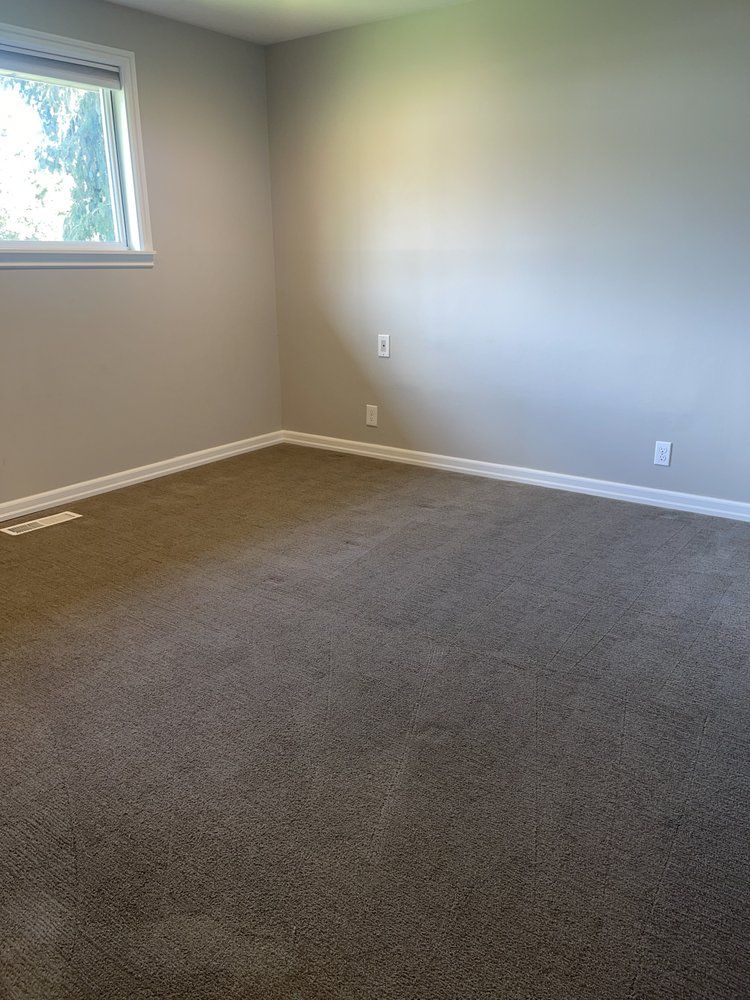 Empty beige carpeted room with light gray walls, a small window, and white baseboards.