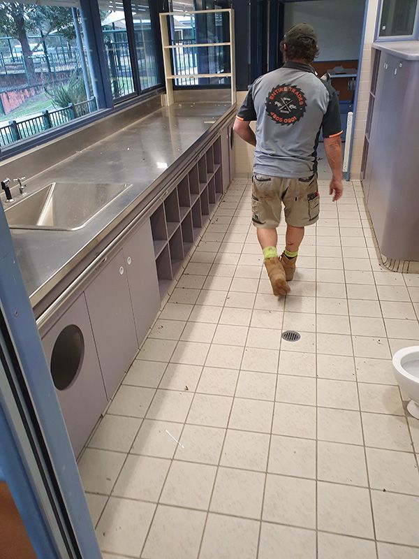 A Worker Man Is Walking Down A Tiled Hallway Towards A Sink And Toilet — Entire Trades in Port Stephens, NSW
