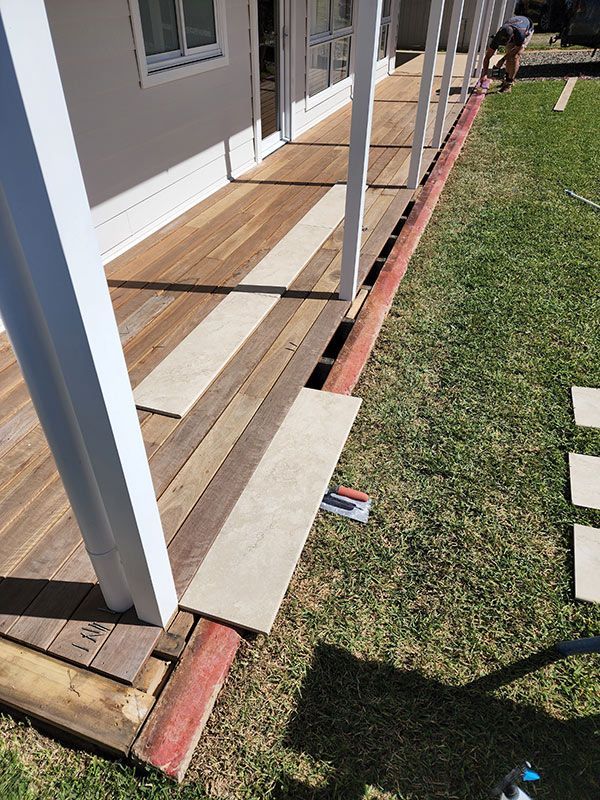 A Man Is Laying Tiles On A Wooden Deck Next To A House — Entire Trades in Singleton, NSW