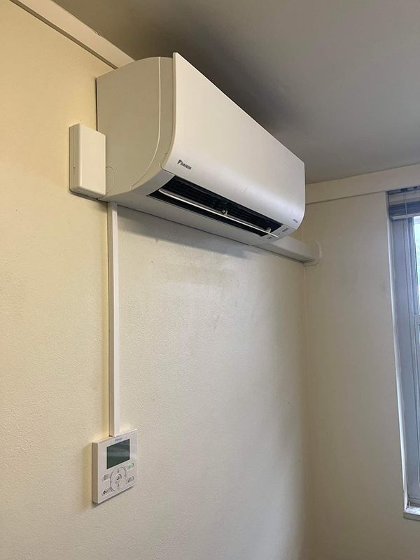 A White Daikin Air Conditioner Is Mounted On A White Wall Next To A Window — Entire Trades in Maitland, NSW