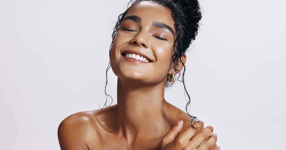 A woman with a towel wrapped around her head is touching her face in front of a mirror.