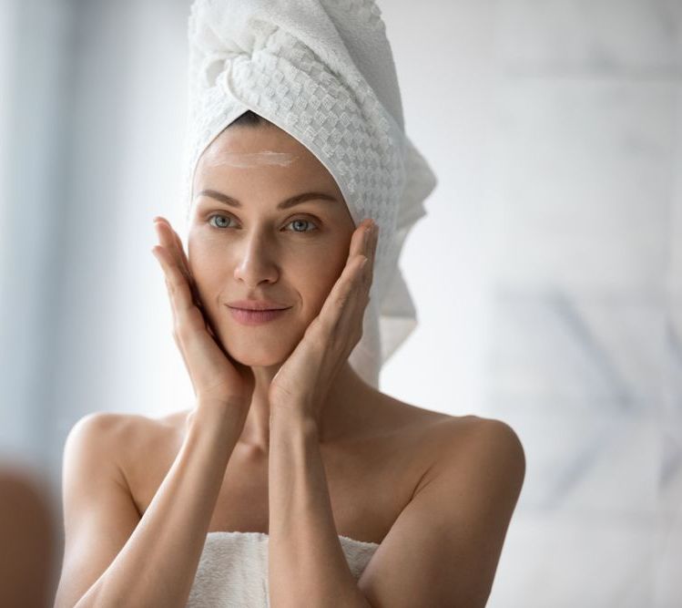 A woman with a towel wrapped around her head is touching her face in front of a mirror.