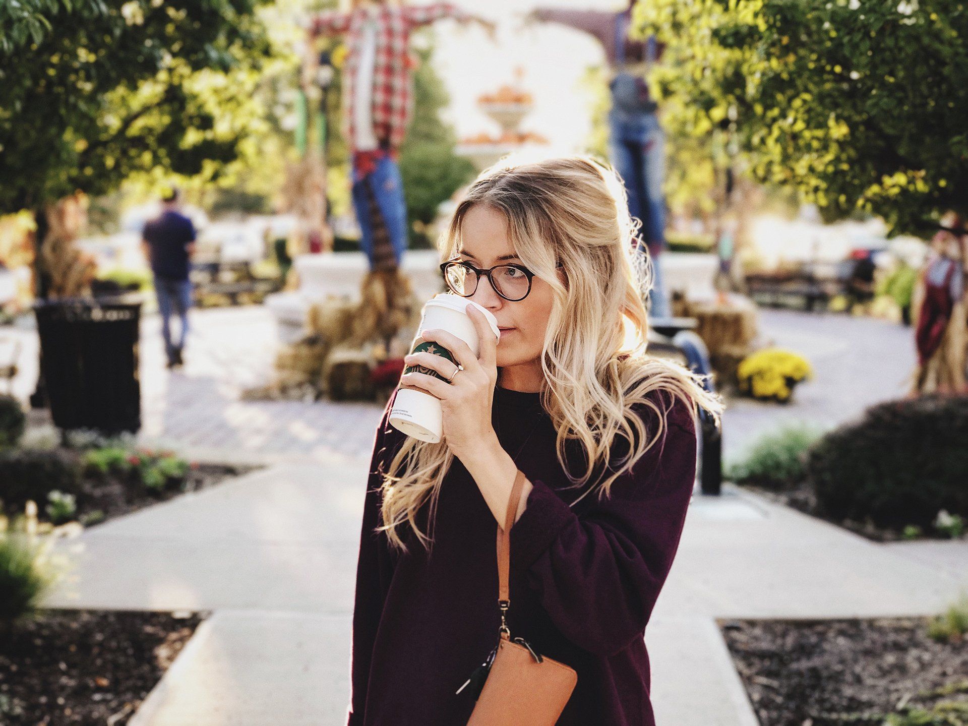 Woman in glasses sips from a coffee cup outdoors; fall setting with scarecrows and foliage.