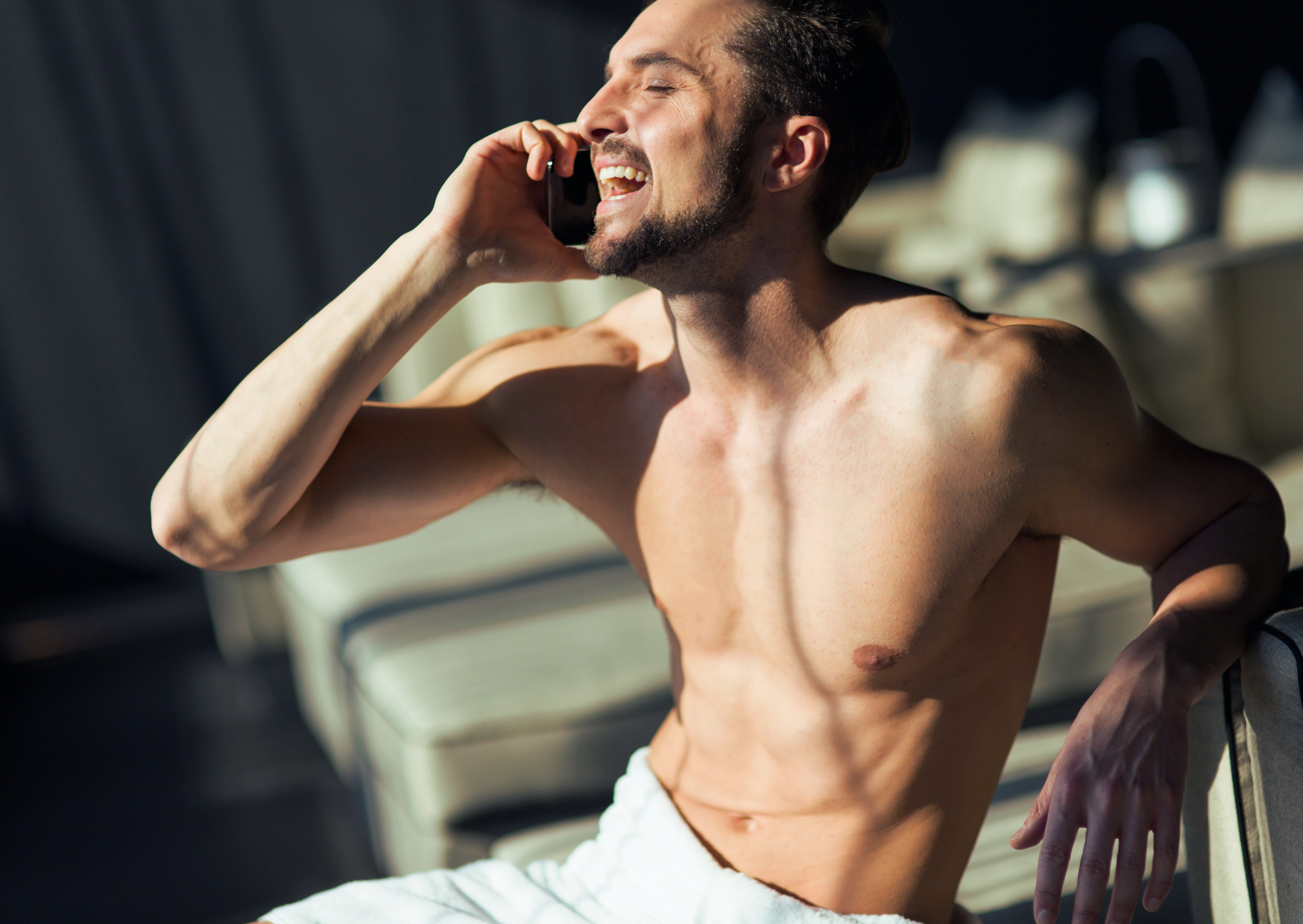 A shirtless man is sitting on a couch talking on a cell phone.