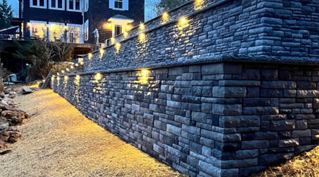 Stone retaining wall with pathway, lit by warm lights at night. House in background.