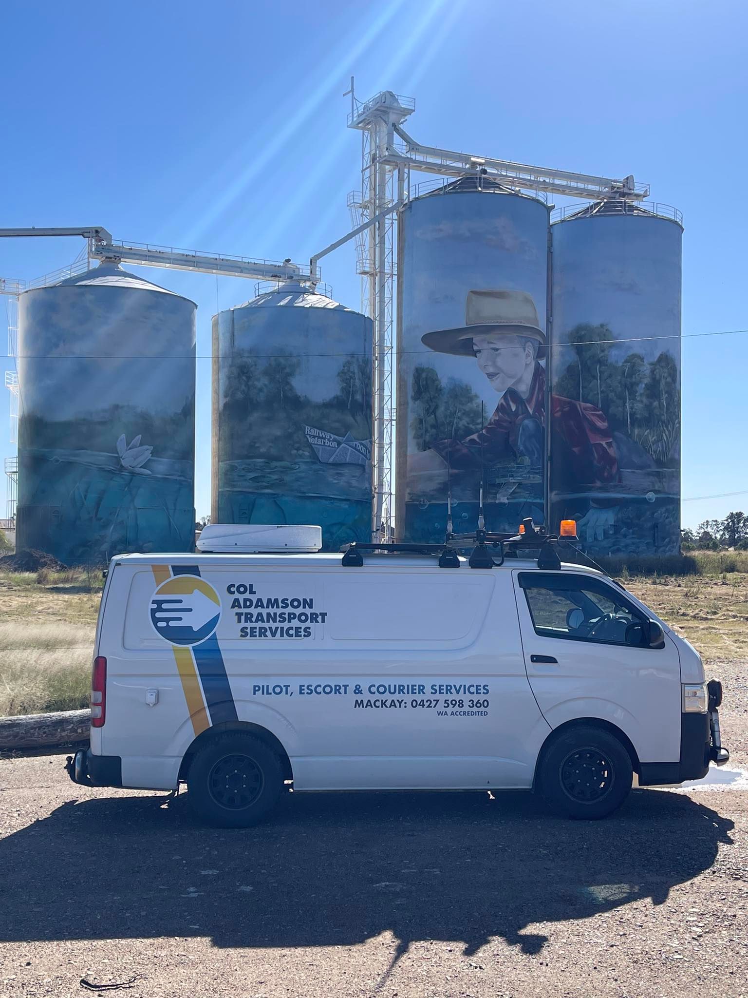 White van parked in front of grain silos with a painted mural; blue sky background — Col Adamson Transport Services in Farleigh, QLD
